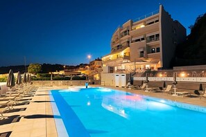 Una piscina con sillas de descanso y sombrillas, un edificio de varios pisos con balcones y un cielo despejado.