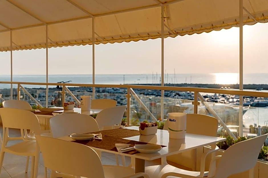 Comedor con sillas y mesas blancas, vista al mar y un paisaje urbano de fondo.