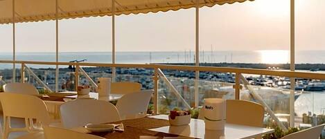 Comedor con sillas y mesas blancas, vista al mar y un paisaje urbano de fondo.