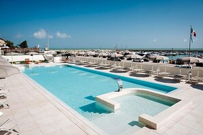 Una piscina con sillas de descanso y sombrillas, una bandera y un puerto deportivo al fondo.