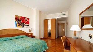 A hotel room with a bed, a desk with a lamp, a chair, a painting on the wall, and a door leading to another room. - Hotel Scogliera (Numana)