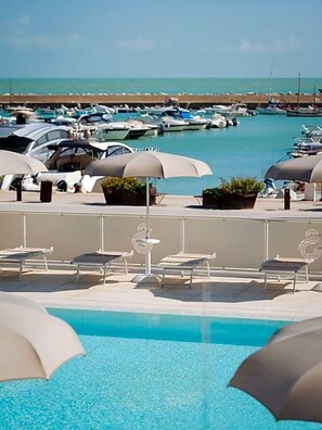 Un área de piscina con sillones de descanso y sombrillas, un puerto deportivo con barcos al fondo y un cielo despejado.