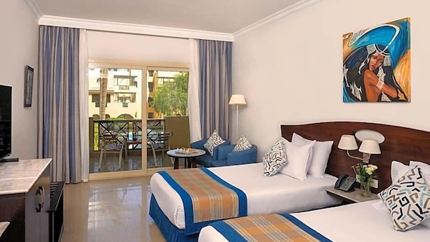 A hotel room with two beds, a TV, a desk, a balcony with a table and chairs, and a painting on the wall. - Stella Gardens Resort Spa Makadi Bay (Makadi Bay)