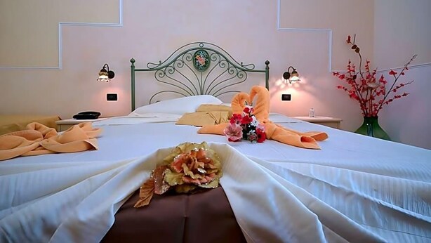A neatly made bed with a heart-shaped towel, a bouquet of flowers, and a vase on the nightstand. - Hotel Ristorante Oasi Da Paolo (San Vito Lo Capo)
