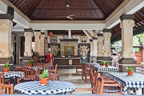 A covered outdoor seating area with tables and chairs, a central counter with a decorative backdrop, and a ceiling fan. - Troppo Zone Puri Rama Resort (Kuta)