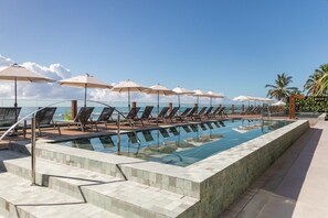 Un espace aménagé au bord de la piscine, avec des chaises longues et des parasols, offrant une vue dégagée sur l’océan, et une piscine carrelée.
