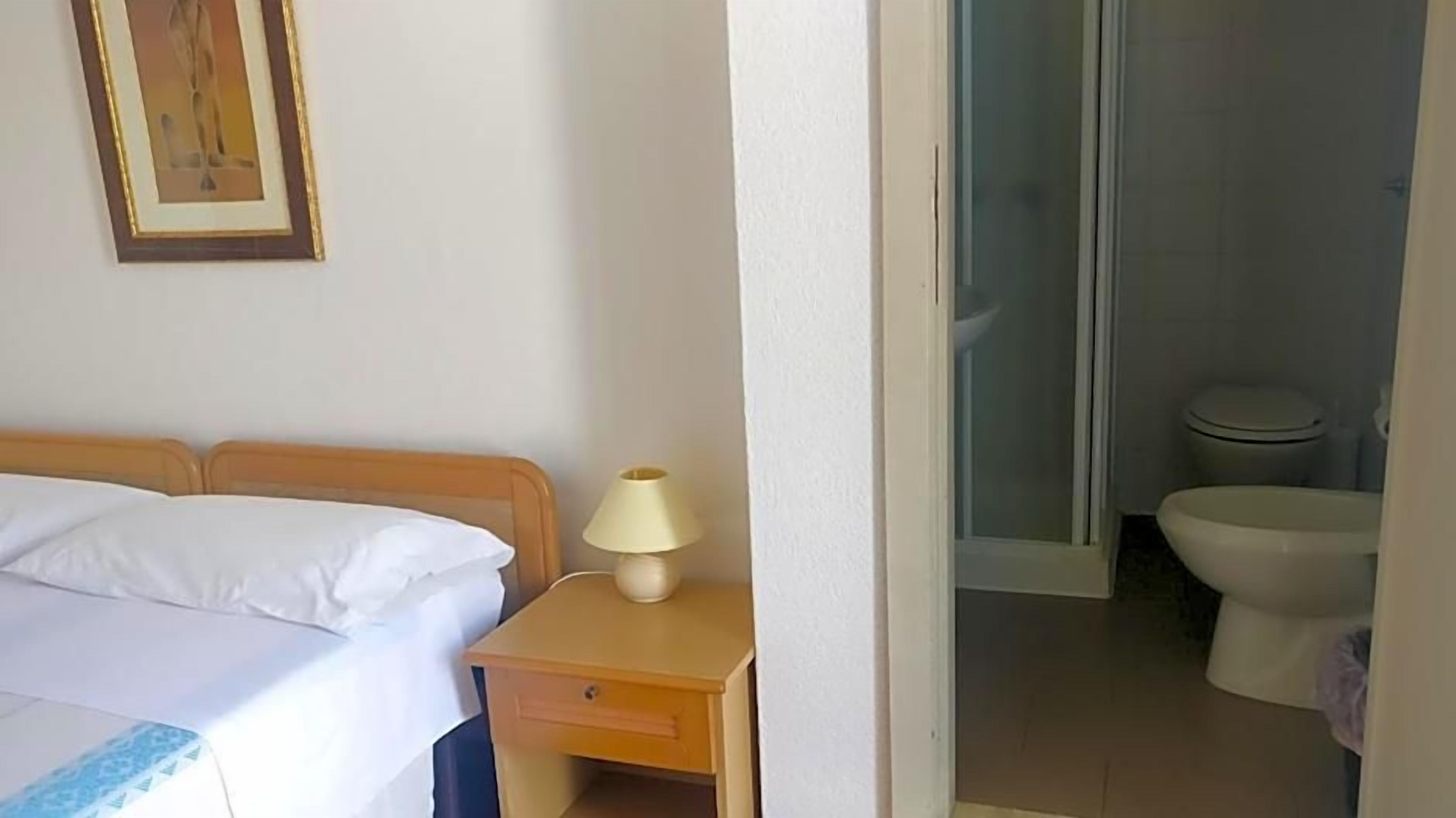 A hotel room with a bed, a nightstand with a lamp, and a bathroom visible through an open door.