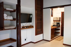 Habitación de hotel con televisión montada en la pared, estantes de madera con toallas, zona de cocina con microondas y cafetera, y suelo de madera.