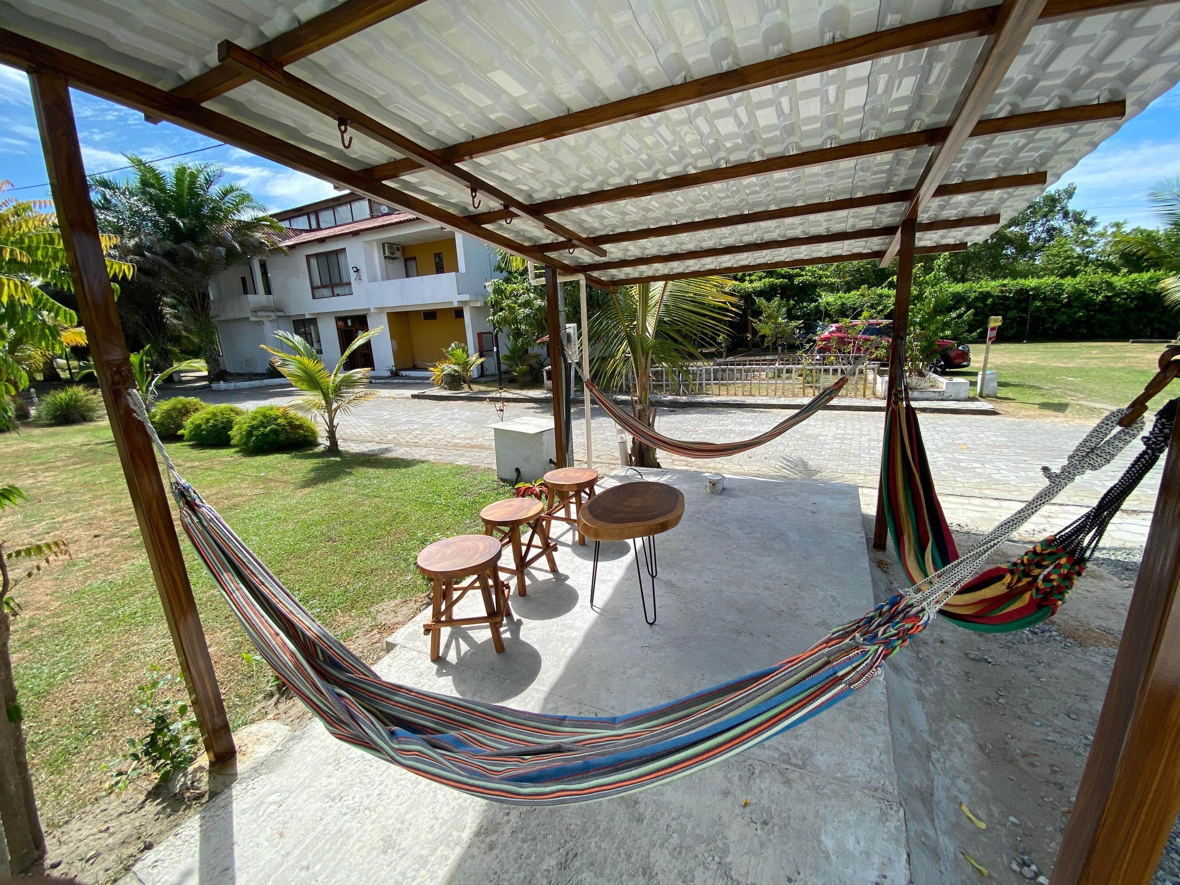 Un espacio de estar al aire libre con techo, que incluye una hamaca, mesas y sillas de madera.