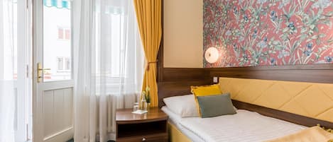 Quarto de hotel com cama, mesa de cabeceira, luminária e janela com cortinas.