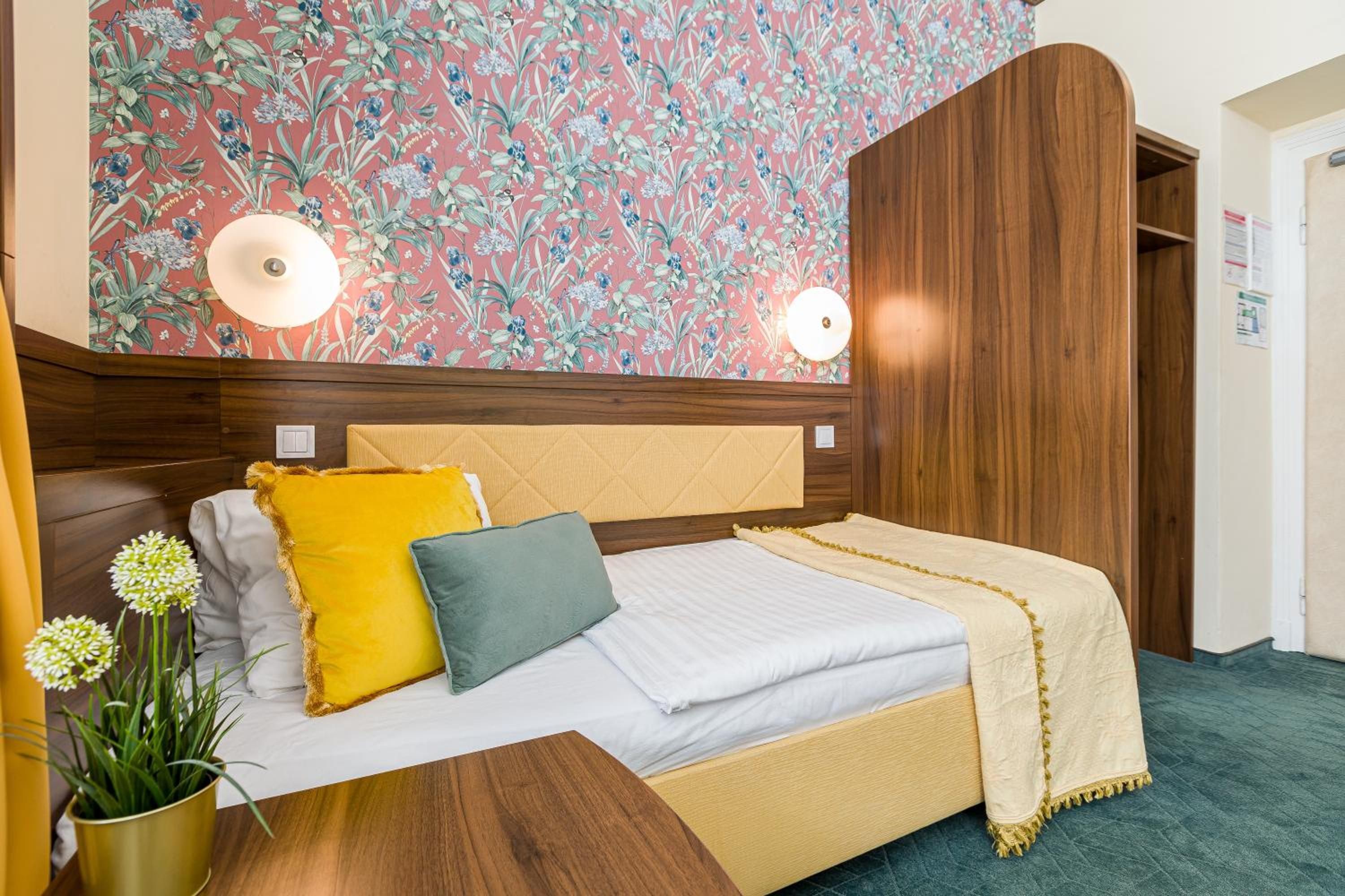 Una habitació d'hotel amb un llit, un panell de fusta al capçal, paper pintat amb motius florals i una catifa verda.