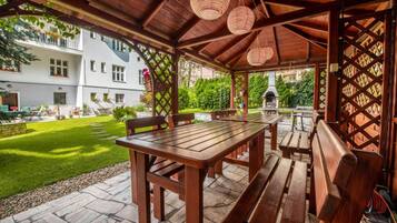 A covered outdoor seating area with a wooden table and benches, stone flooring, and a view of a building and garden.