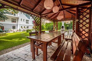 Uma área de estar coberta ao ar livre, com uma mesa de madeira e bancos, piso de pedra e vista para um edifício e jardim.