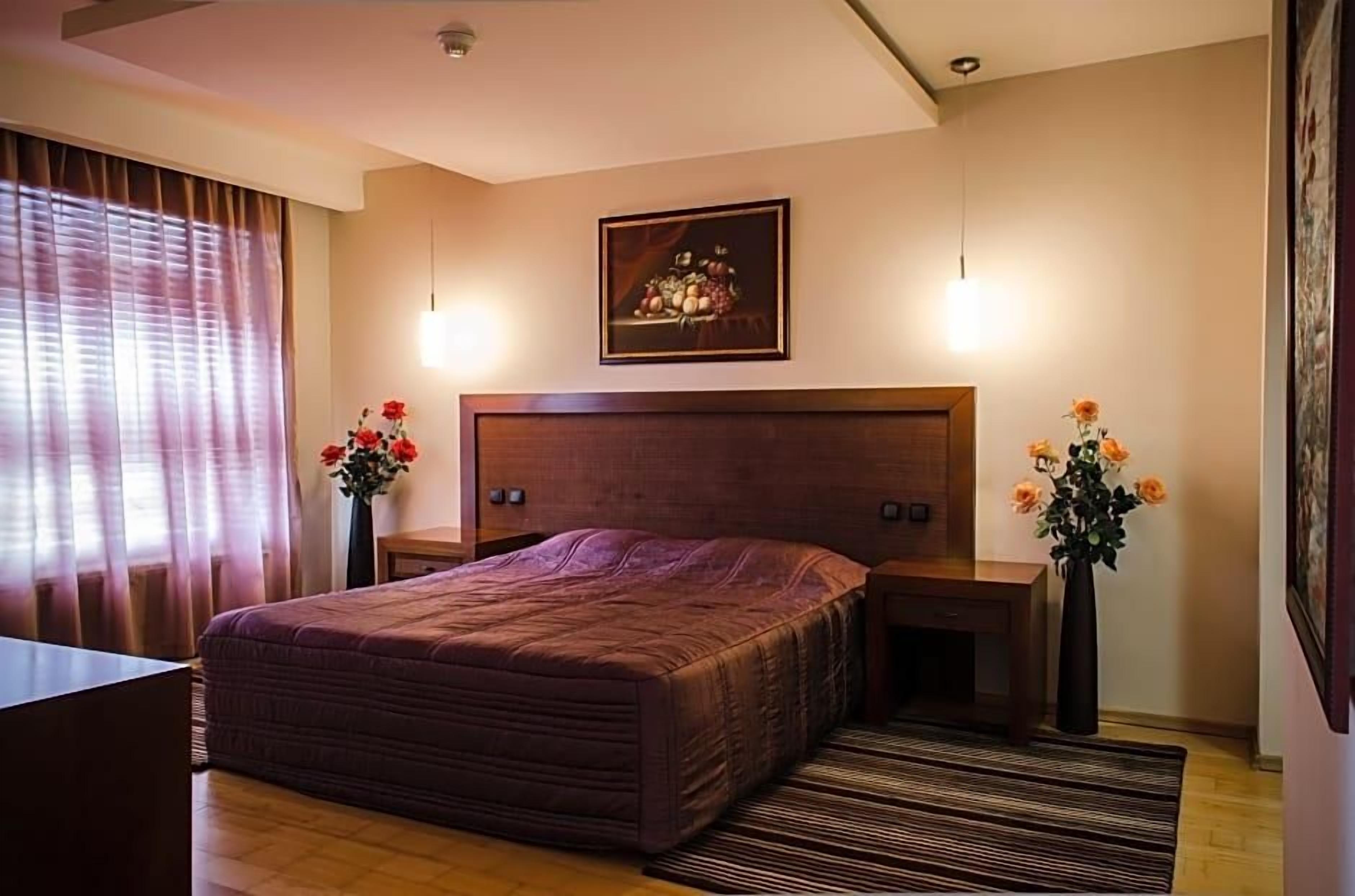 Un dormitorio con una cama grande, mesitas de noche, una obra de arte enmarcada sobre la cama y una ventana con cortinas.