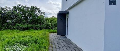 A modern building with a paved pathway leading to a dark entrance door, surrounded by lush greenery.