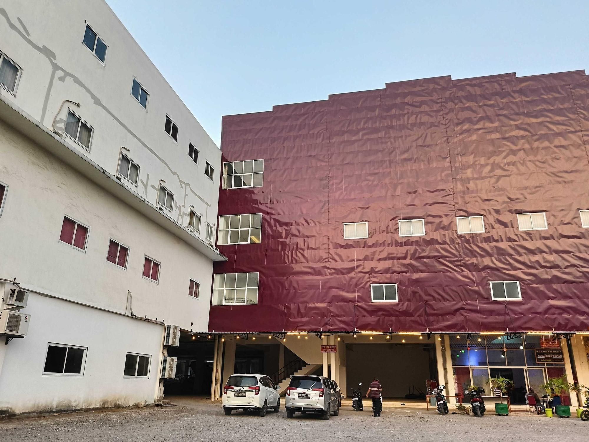 A multi-story building with a red facade and a white building adjacent to it. There are parked cars and a person walking near the entrance.