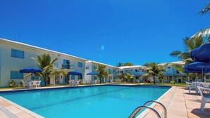 Una piscina con sillas de descanso y sombrillas, rodeada de edificios con balcones y palmeras.