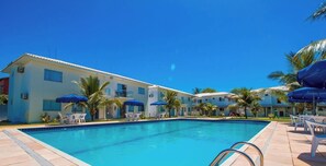 A swimming pool with lounge chairs and umbrellas, surrounded by buildings with balconies and palm trees. - Porto Luar Apart Hotel (Porto Seguro)