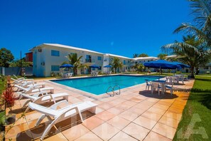 A swimming pool with lounge chairs and umbrellas, a building with balconies, and a clear blue sky. - Porto Luar Apart Hotel (Porto Seguro)