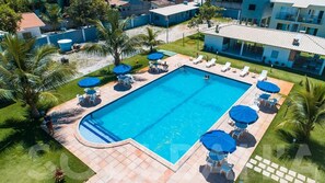 Una piscina rectangular con un borde de baldosas, rodeada de sillas de descanso y sombrillas, ubicada en una zona ajardinada con palmeras y un edificio al fondo.