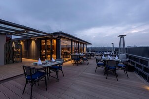 Un área de comedor al aire libre con mesas y sillas, una sección cubierta con paredes de vidrio y vistas al horizonte de la ciudad.