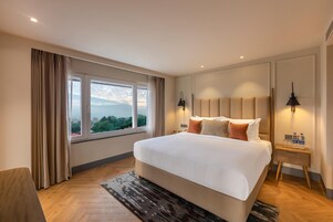 Una habitación de hotel con una cama grande, una mesita de noche con lámpara, vistas de la ciudad a través de la ventana y suelo de madera.