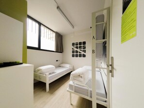 A dormitory room with two single beds, a window, and a door with a bed on the other side. - Room018bcn (Barcelona)