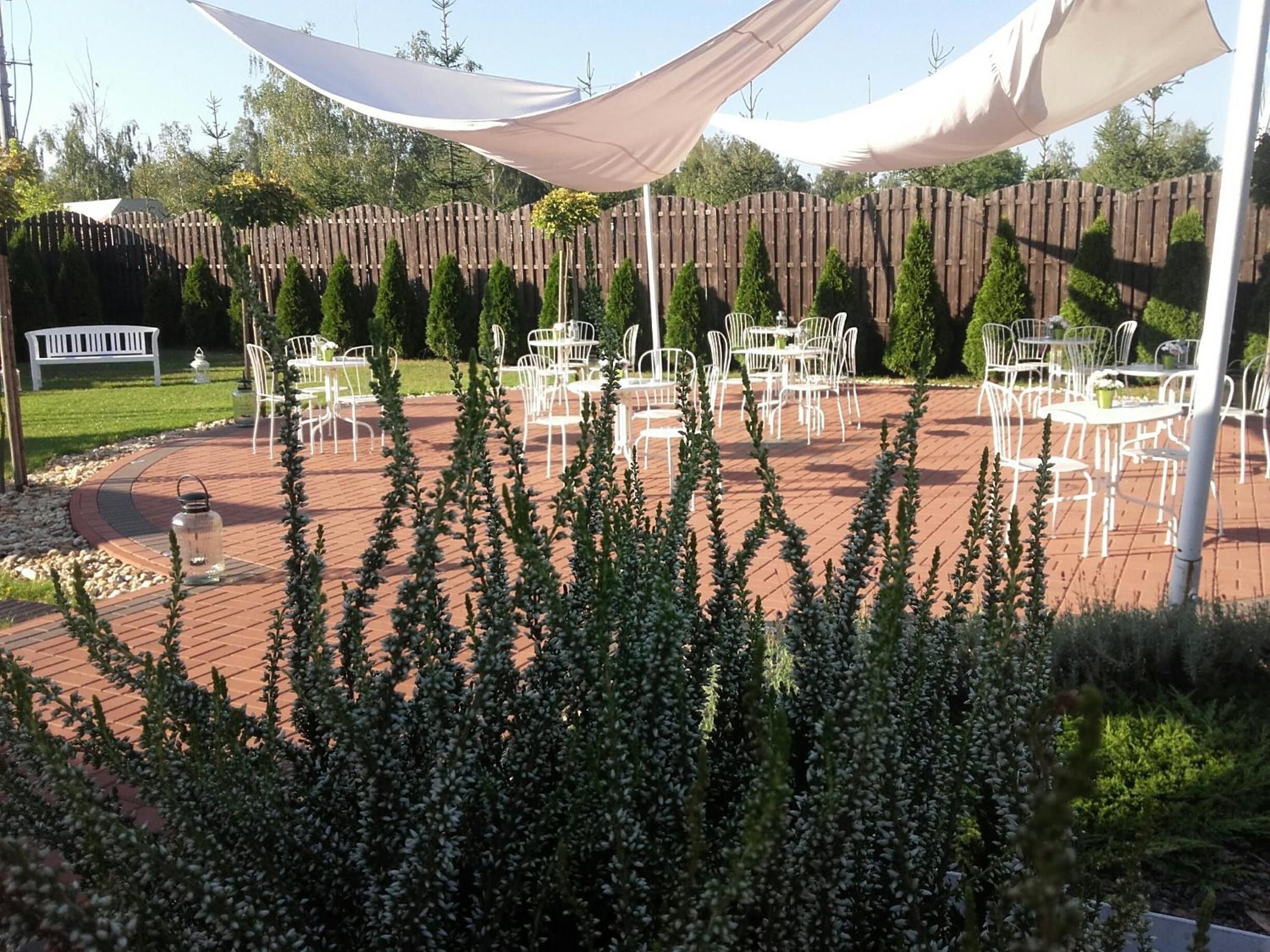 Eine Terrasse im Freien mit weißen Metallstühlen und -tischen, umgeben von einem Holzzaun und viel Grün.