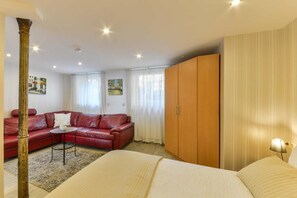 Habitación de hotel con un sofá de cuero rojo, una mesita redonda pequeña, una cama con edredón blanco, armario de madera y dos cuadros en la pared.