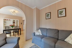 Uma sala de estar com um sofá cinzento, uma mesa de jantar com cadeiras e um candelabro.
