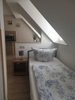 Quarto com teto inclinado, cama com colcha florida, mesa de cabeceira com abajur, uma pequena planta em vaso e vista para uma cozinha adjacente.
