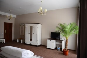Habitación de hotel con cama, televisor, armario, escritorio, silla, pequeña mesa, banco, planta en maceta y candelabro.