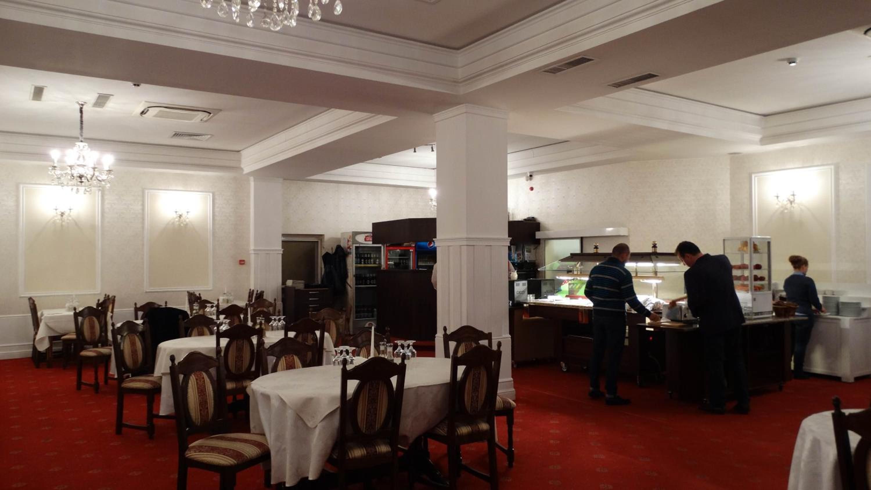 Un comedor de hotel con varias mesas y sillas, una zona de bar con personal y un suelo de moqueta roja.