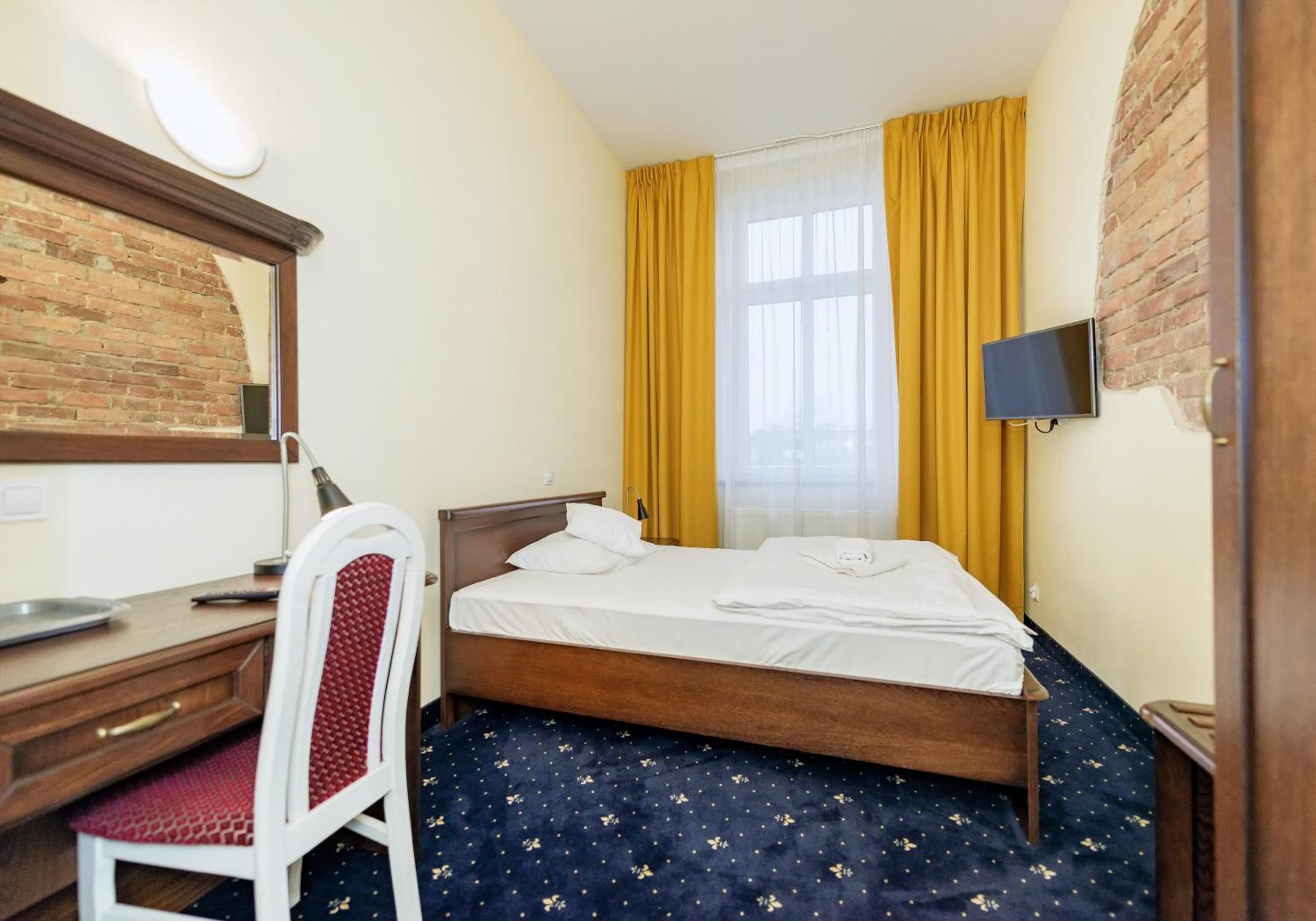 A hotel room with a wooden bed, a desk with a chair, a flat-screen TV mounted on the wall, and a window with yellow curtains.