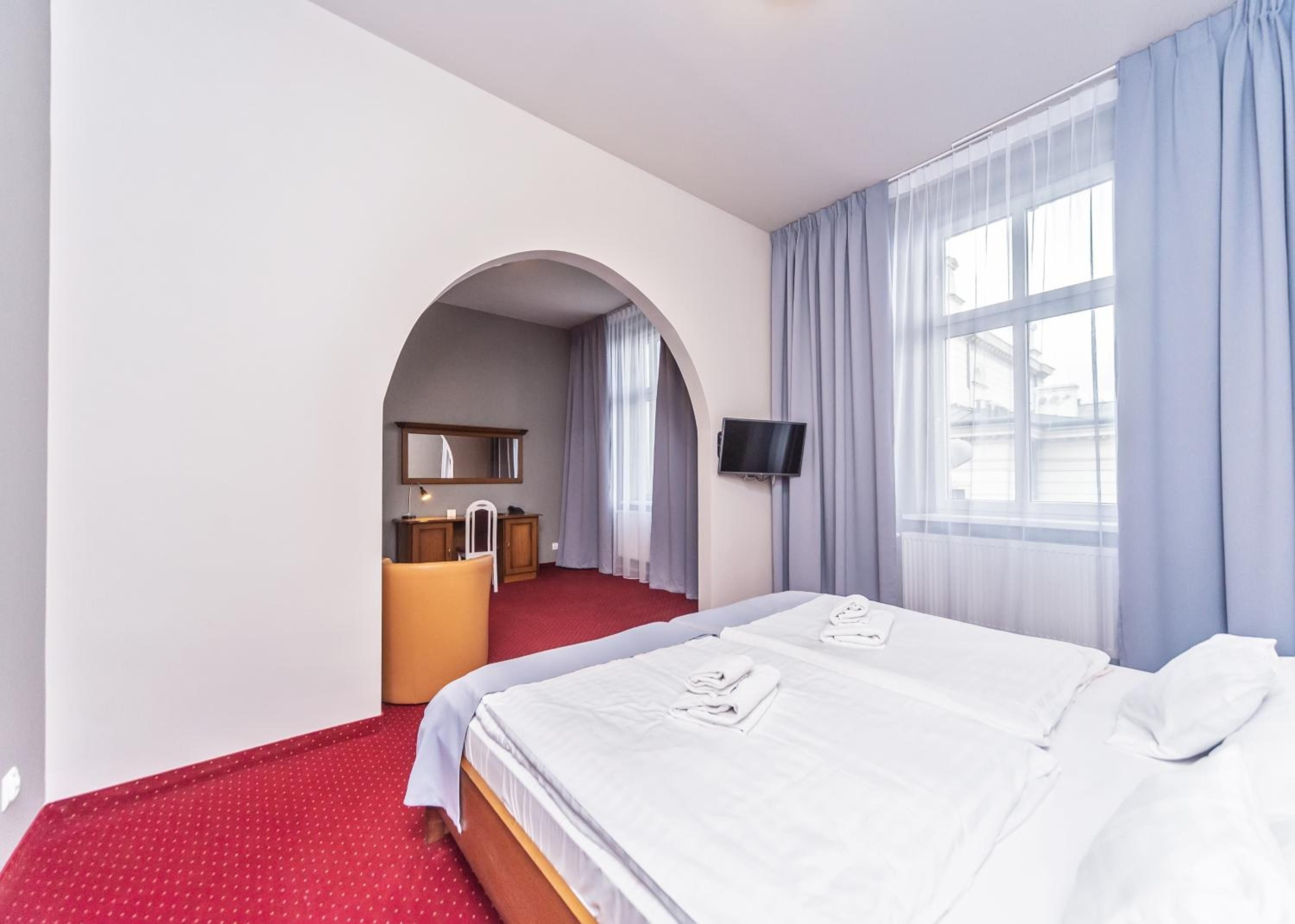 Une chambre d’hôtel comprenant un lit, un bureau avec une chaise, une télévision et une fenêtre avec des rideaux.