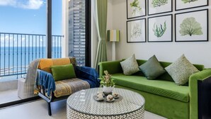 Un balcon avec vue sur la mer, un canapé vert agrémenté de coussins à motifs, un fauteuil en osier avec une couverture, et une table ronde avec une théière et des tasses.