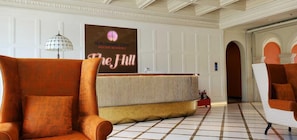 Uma área de recepção com uma grande placa que diz 'The Hill Hillside Residence', um balcão de atendimento e duas poltronas grandes.