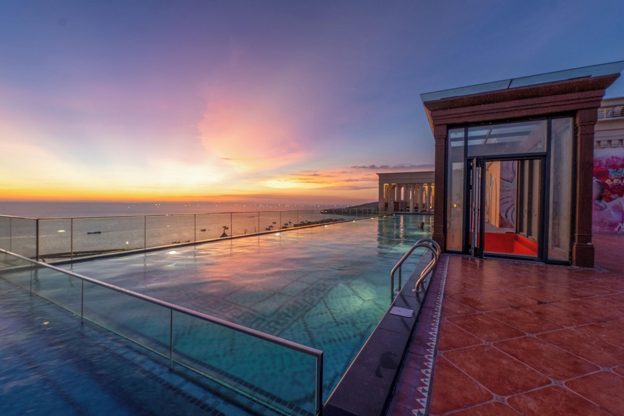 A rooftop infinity pool with a glass barrier, a red-tiled floor, and a view of the sunset over the ocean.