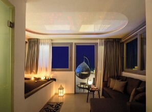 Ein modernes Wohnzimmer mit Sofa, Hängekorbschaukel, kleinem Tisch mit Kerzen und Blick auf den Nachthimmel durch ein Fenster.