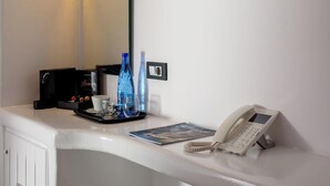 A hotel room with a white desk, a mirror, a black coffee machine, a phone, and two blue water bottles. - Tataki Family Hotel (Santorini)