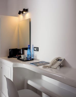 Une chambre d’hôtel avec un bureau blanc, un miroir, une machine à café noire, un téléphone et deux bouteilles d’eau bleues.