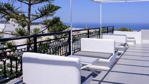 A balcony with white furniture, a metal railing, and a view of the sea and buildings. - Tataki Family Hotel (Santorini)