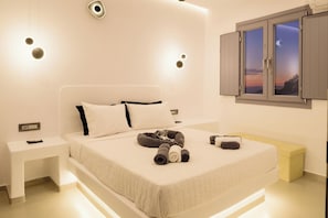 A modern bedroom with a bed, bedside tables, a bench, and a window with a view. - Tataki Family Hotel (Santorini)