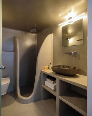 A bathroom with a curved shower, a toilet, a sink with a stone basin, and a mirror with a light above it. - Tataki Family Hotel (Santorini)