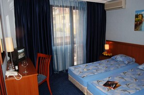 A hotel room with a bed, a desk with a telephone and glasses, a chair, a nightstand with a lamp, and a painting on the wall. - Hotel Kibella (Sofia)