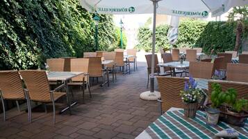 An outdoor patio area with wicker furniture, tables, and umbrellas, surrounded by greenery.