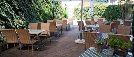 Un espacio al aire libre con patio, mobiliario de mimbre, mesas y sombrillas, rodeado de vegetación.