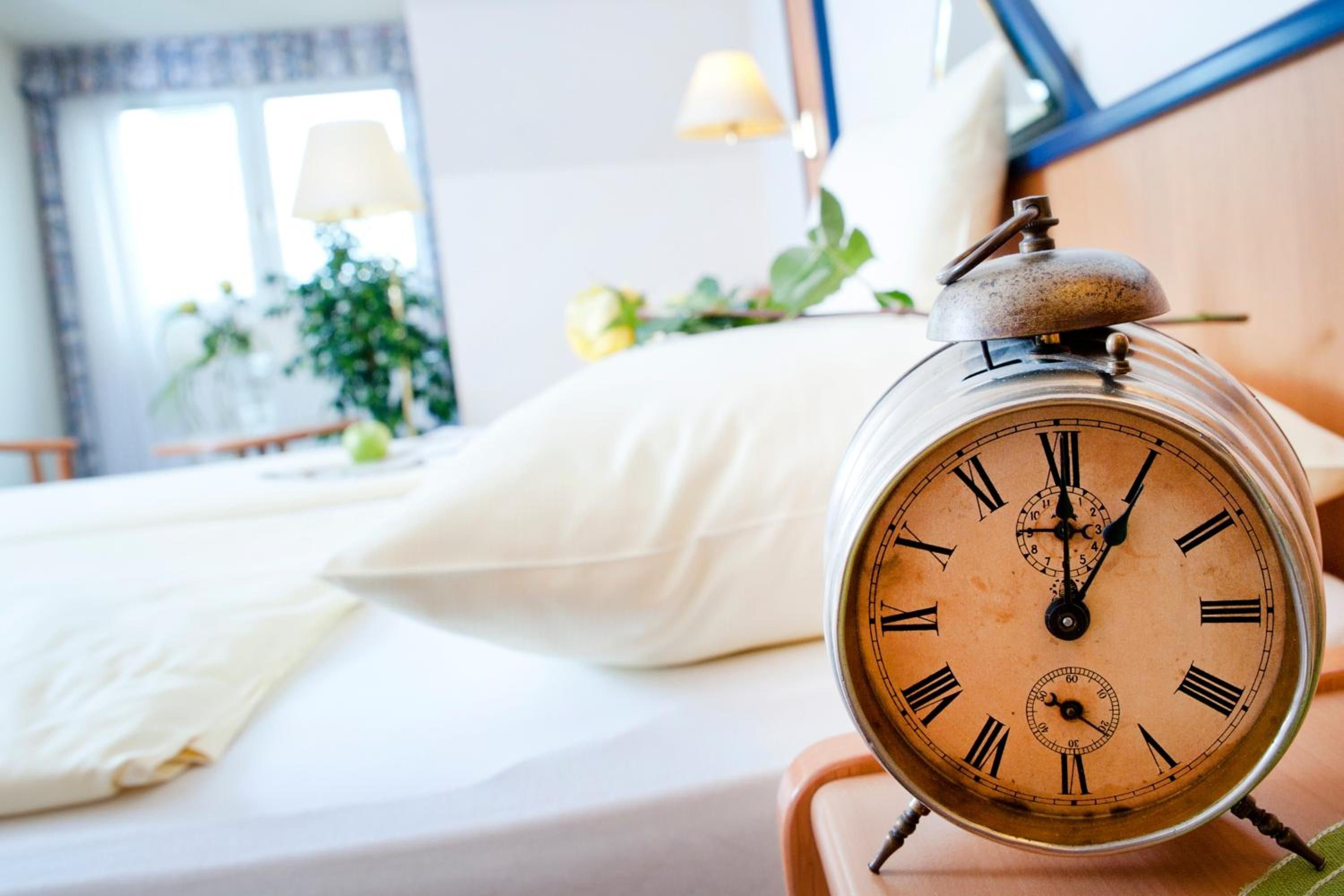 Eine alte Weckuhr auf einem Nachttisch in einem Hotelzimmer, mit einem Bett, Kissen und einem Fenster mit Vorhängen.