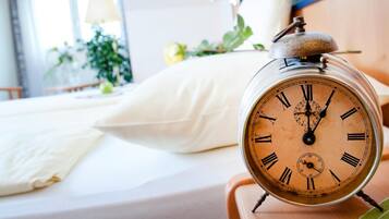 Eine alte Weckuhr auf einem Nachttisch in einem Hotelzimmer, mit einem Bett, Kissen und einem Fenster mit Vorhängen.