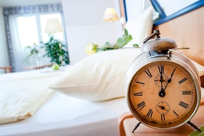 Un reloj despertador antiguo sobre una mesita de noche en una habitación de hotel, con una cama, almohadas y una ventana con cortinas.
